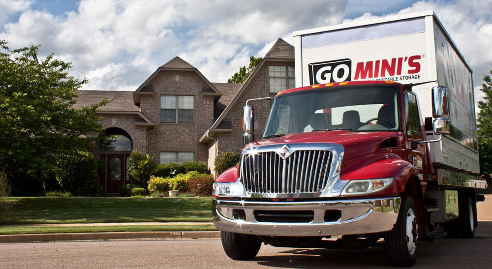 A flat-bed truck carrying a Go Mini's portable storage container parked in front of a house