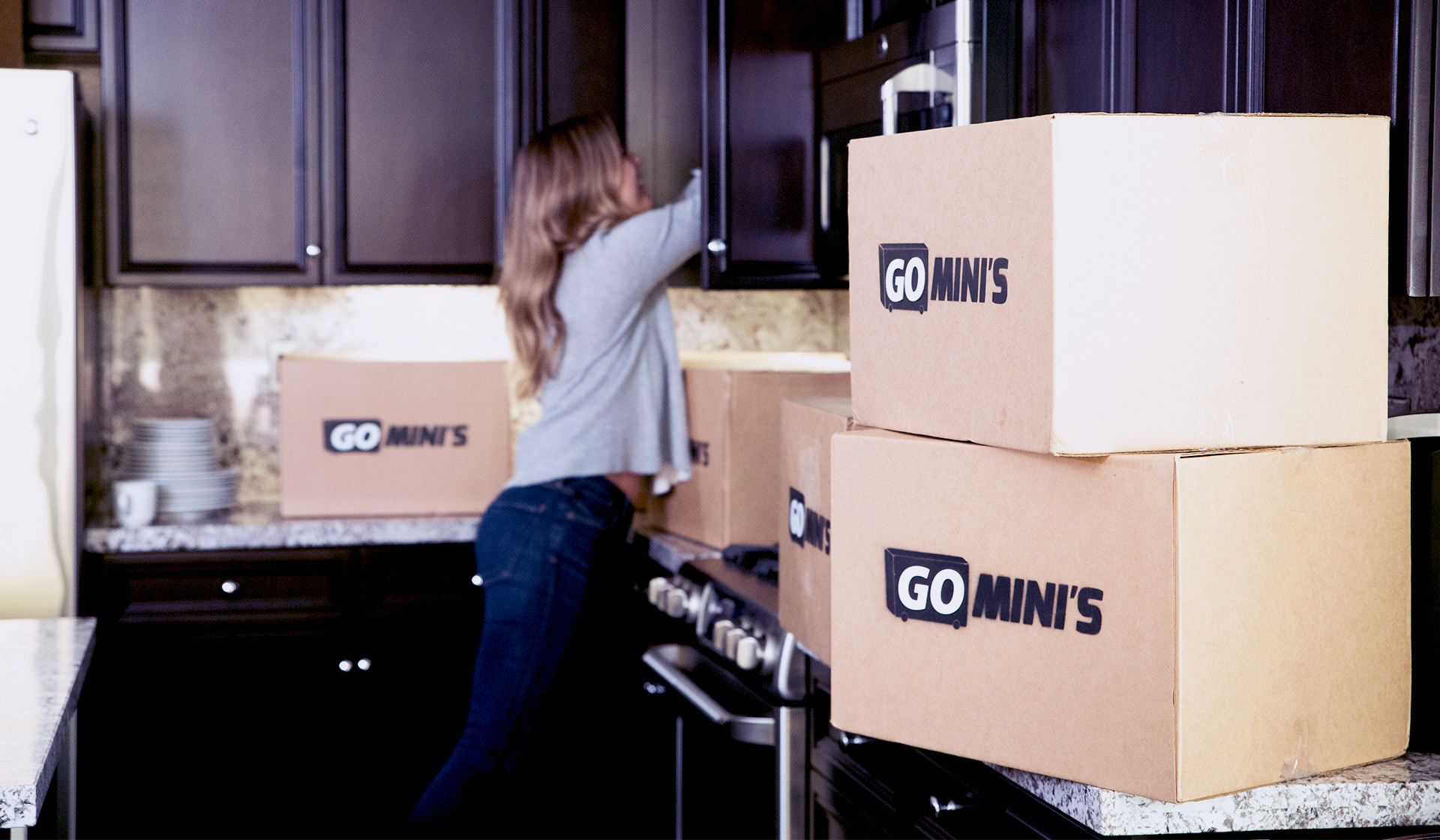 A woman reaches into a kitchen cupboard while cardboard Go Mini's moving boxes are on the counter